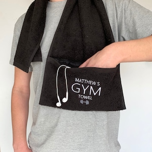 Könnte beinhalten: Schwarzes Sporttuch mit einer Tasche und einem Reißverschluss. Das Handtuch trägt den Text "Matthew's Gym Towel" und ein gesticktes Hantel-Symbol.