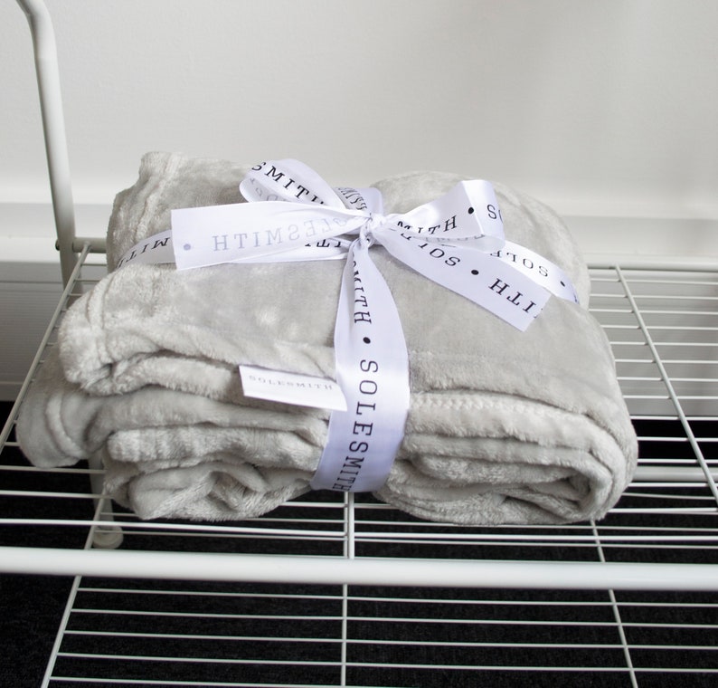 May include: A gray plush throw blanket tied with a white satin ribbon. The ribbon has the text "SOLESMITH" printed on it.