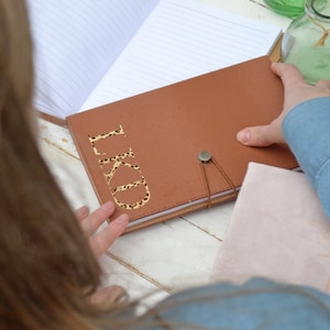 May include: A brown leather journal with a leopard print monogram "LKD" on the front cover. The journal has a strap closure and a metal button.