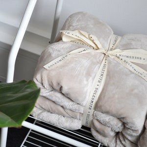 May include: A soft, beige throw blanket folded and tied with a cream-colored ribbon. The ribbon has the text "SOLESMITH" printed on it.