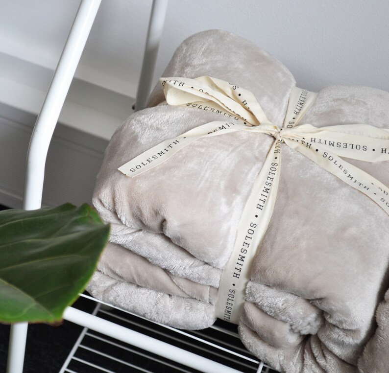 May include: A soft, beige throw blanket folded and tied with a cream-colored ribbon. The ribbon has the text "SOLESMITH" printed on it.