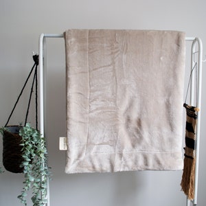 May include: A beige faux fur throw blanket draped over a white metal rack. The blanket has a subtle sheen and a soft, plush texture.