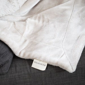 May include: A white sherpa throw blanket with a white tag that says "SOLESMITH" on a dark gray surface.