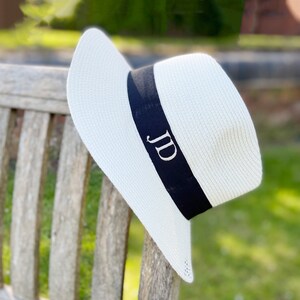 May include: A white sun hat with a wide brim and a black band. The band features the embroidered initials "JD" in white. The hat is made of woven material and is resting on a wooden bench.