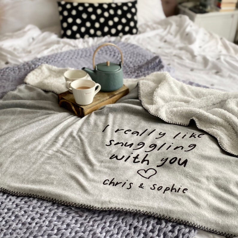 May include: A gray fleece blanket with the text "I really like snuggling with you" and a heart symbol. The text "chris & sophie" is below the heart. Two mugs and a teapot are on a wooden tray in the background.