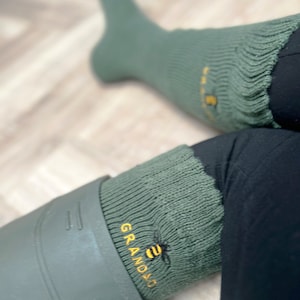 Può includere: Calze di lana verde con un logo a forma di ape e il testo "GRANDAD" ricamato sul lato. Le calze sono indossate con pantaloni neri e stivali di gomma verdi.