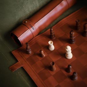 Könnte beinhalten: Ein braunes Schachspiel aus Leder mit einer aufgerollten Tragetasche. Das Schachbrett ist ausgeklappt und zeigt ein laufendes Spiel mit dunklen und hellen Figuren. Auf der Tasche steht der Name "JAMES".