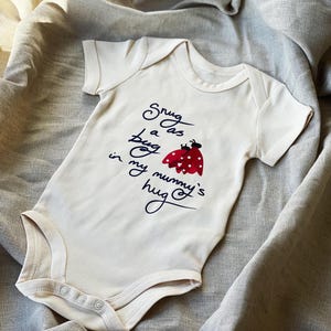 Op de afbeelding: Witte baby romper met een rode lieveheersbeestje afbeelding en de tekst "Snug as a bug in my mummy's hug".