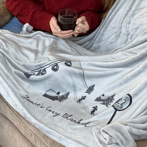 Può includere: Una morbida coperta in pile grigia con un design a tema invernale. La coperta presenta una seggiovia, un chalet e un cartello con la scritta "Crest 22 Vercland". Il testo "Jamie's Cozy Blanket" è scritto in corsivo sulla coperta.