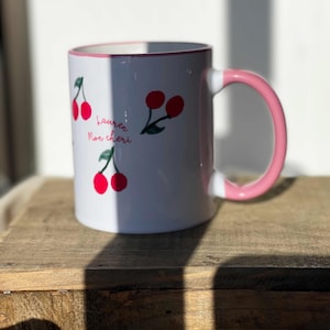 May include: White ceramic mug with a pink handle and rim, adorned with red cherry illustrations and the text "Lauren Mon cheri". The mug is set on a wooden surface, with sunlight casting shadows.