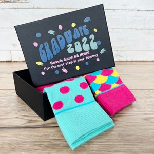 Puede incluir: Una caja de regalo negra con un diseño colorido "Graduate 2022" y el texto "Hannah Smith BA Hons Para el siguiente paso en tu viaje". La caja está abierta y contiene dos pares de calcetines coloridos, uno con lunares rosas sobre un fondo turquesa y el otro con un patrón geométrico rosa, amarillo y azul.