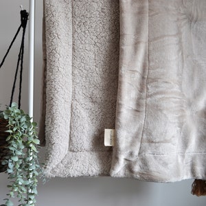 May include: A beige throw blanket with a soft, fuzzy texture on one side and a smooth, velvety texture on the other side. The blanket has a white tag with the text "Made with Love" on it.