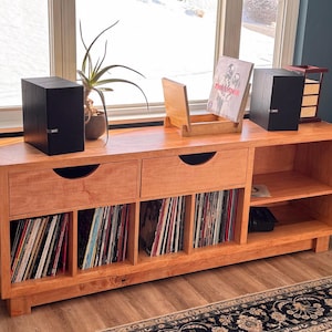 May include: A wooden media console with two drawers and open storage for vinyl records. The console is made of light-colored wood and has a mid-century modern design. Two black speakers and a record stand are on top.
