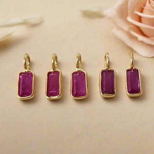 May include: Five gold-coloured rectangular pendants with magenta-coloured stones. Each pendant has a small loop at the top. A pink rose is in the background.
