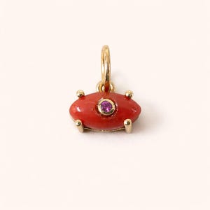 Peut inclure: Un charm doré avec une pierre centrale rouge de forme ovale et une petite pierre violette au centre. Le charm possède une boucle pour l'accrocher et quatre griffes dorées.