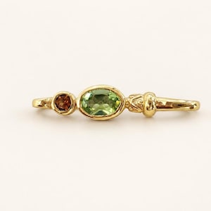 Könnte beinhalten: Ein Goldring mit einem zentralen oval geschliffenen grünen Edelstein, einem kleineren rund geschliffenen braunen Edelstein auf der linken Seite und einem dekorativen, strukturierten Metalldetail auf der rechten Seite des polierten Goldrings.