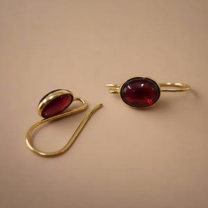 Può includere: Un paio di orecchini color oro con una pietra rosso scuro a forma ovale. Un orecchino ha un design curvo, l'altro è un semplice cerchio. Gli orecchini sono su uno sfondo neutro.