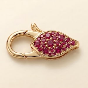 May include: A gold jewelry clasp featuring a teardrop-shaped section encrusted with numerous small, round, deep red rubies. The clasp has a polished metallic finish and a spring-loaded mechanism for securing necklaces or bracelets.
