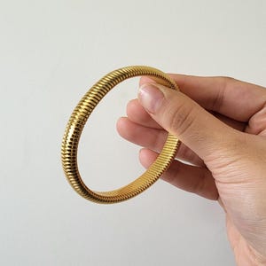 Pode incluir: Uma pulseira dourada e canelada. A pulseira tem um design flexível e enrolado. A joia é segurada contra um fundo branco.