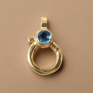 Puede incluir: Un colgante dorado con un diseño de cierre redondo y abisagrado. El colgante presenta una piedra preciosa redonda de color azul claro engastada en el centro. El cierre está unido a un pequeño lazo en la parte superior.
