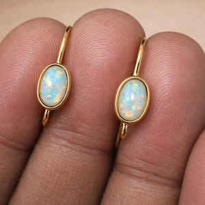 Può includere: Un paio di orecchini color oro, ciascuno con una pietra di opale ovale. Gli opali mostrano un gioco di colori con lampi iridescenti di blu, verde e rosa. Gli orecchini sono tenuti da un dito.