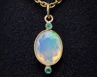 May include: A gold necklace featuring an oval, faceted opal pendant with a gold bezel. Two small, round, green gemstones are set above and below the opal. The chain is gold and the background is black.
