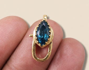 May include: A gold-colored clasp with a marquise-cut, teal-colored gemstone. The clasp is designed with a decorative, scalloped bezel around the gem and a curved, oval-shaped base. The clasp is likely for jewelry.