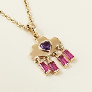 May include: Gold-toned necklace featuring a cloud-shaped pendant with a heart-shaped purple gemstone and three dangling rectangular pink gemstones. The chain is a delicate link style.
