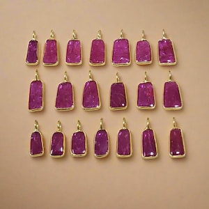 May include: A selection of rectangular pendants featuring magenta gemstones set in gold-coloured metal frames. The pendants are arranged in rows, displaying their polished surfaces and gold loops against a neutral backdrop.