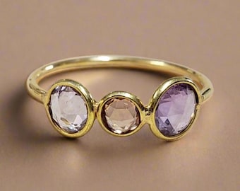May include: A gold-toned ring featuring three oval-cut gemstones in shades of lavender, peach, and purple. The ring's band is a simple, polished design, showcasing the colorful stones. The gemstones are set in a classic, elegant style.