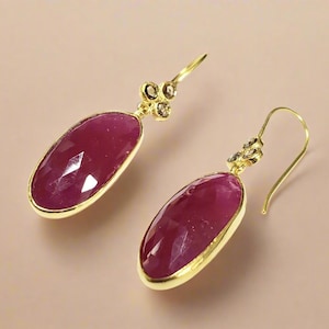 May include: A pair of gold earrings featuring oval-shaped pink gemstones and small clear gemstones. The earrings have a simple, elegant design.
