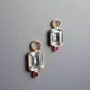 Op de afbeelding: Twee gouden bedels met heldere rechthoekige stenen en kleine roze stenen die aan een gouden lus hangen.
