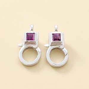 May include: A pair of silver-toned lobster claw clasps, each featuring a square-cut, deep magenta gemstone set into the top rectangular housing. The metal components have a polished, reflective finish with a functional spring-loaded gate mechanism.