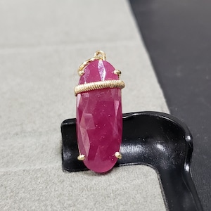 May include: A pink gemstone pendant with a gold wire wrap. The pendant is oval shaped and has a faceted design.