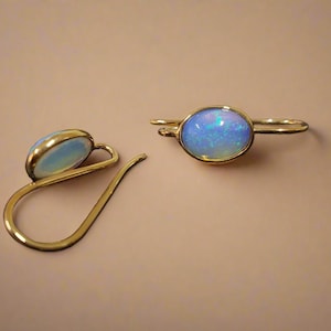May include: A pair of gold-coloured earrings featuring oval opals. The opals display a blue and white iridescent sheen. The earrings are set in gold-coloured metal and have a curved design.