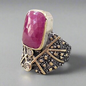 May include: A silver and gold ring with a large, faceted, pink ruby gemstone. The ring has a detailed, textured band with gold accents.