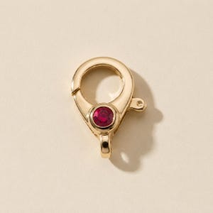 Puede incluir: Un cierre de mosquetón dorado con una piedra preciosa circular de color rojo intenso y tallada en facetas, engastada en el centro del mecanismo. El cierre tiene un acabado liso y pulido, e incluye una pequeña anilla en la parte inferior.