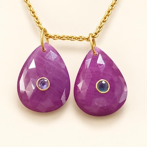 May include: A gold chain necklace featuring two faceted, teardrop-shaped purple gemstone pendants. Each pendant is set with a small, round, blue stone in a gold bezel at its center, suspended from the chain by gold jump rings.
