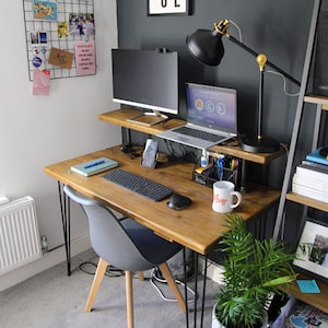 Compact home office desk with a natural wood top and black hairpin legs, featuring a raised shelf holding two monitors and a desk lamp. The workspace includes a grey chair with wooden legs, a keyboard, mouse, notebooks, and various office accessories. A wire memo board with photos is mounted on the white wall, and a shelving unit with books and decor stands on the right. A small green plant sits on the floor in the foreground.