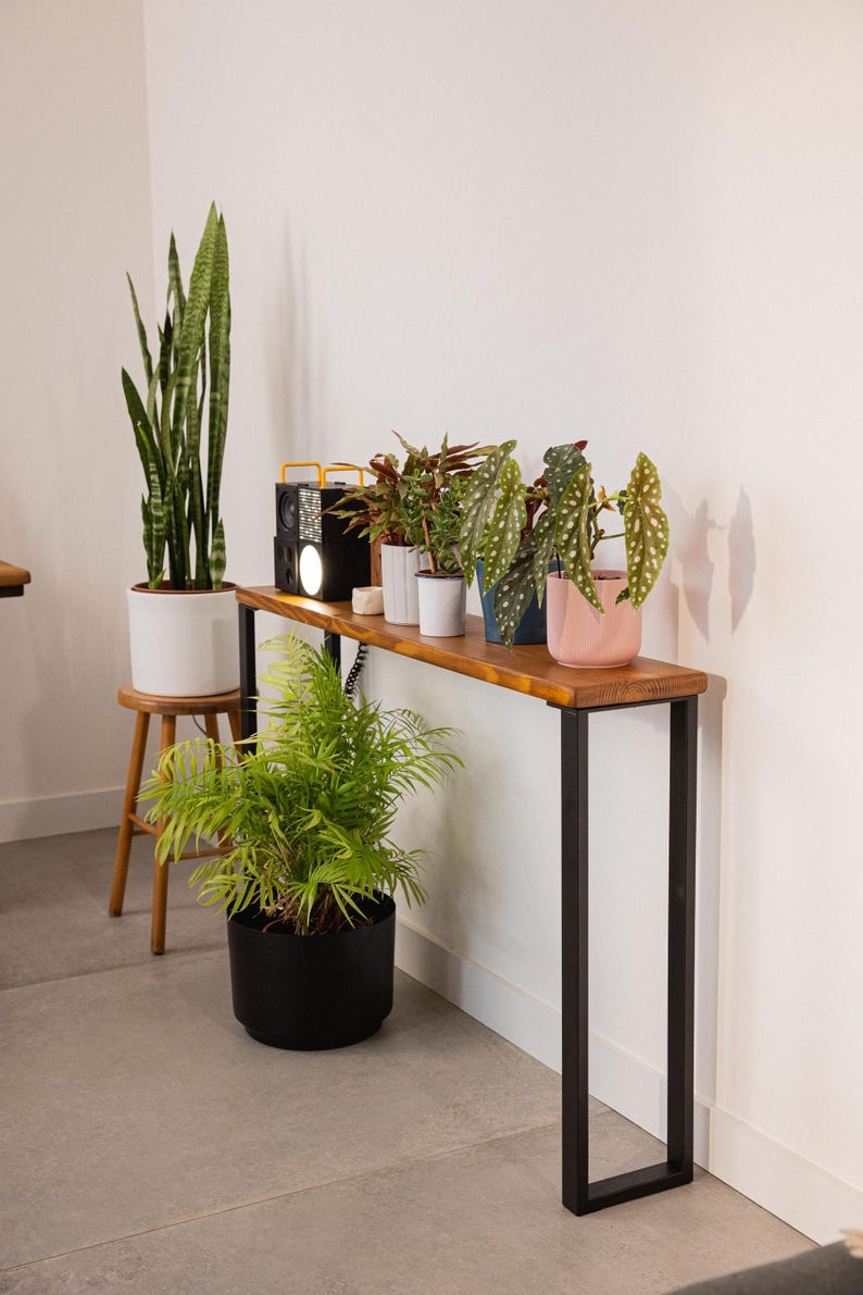 Rustic wooden console table with black steel legs, positioned above a white radiator in a hallway. The table is styled with a potted plant, decorative bowl, and small dish, showcasing its slim design suitable for narrow spaces.