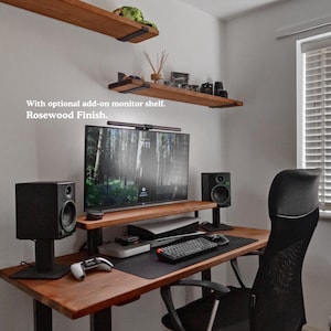 Solid wooden desk with black steel legs, featuring a monitor shelf for a large screen. The desk is styled with studio monitors, a gaming console and a large keyboard mat. The desk is placed in a minimalist room with light off white coloured walls. There are two matching wooden shelves on the wall, above the desk.