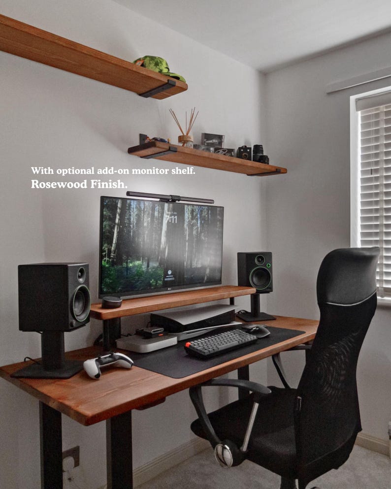 Solid wooden desk with black steel legs, featuring a monitor shelf for a large screen. The desk is styled with studio monitors, a gaming console and a large keyboard mat. The desk is placed in a minimalist room with light off white coloured walls. There are two matching wooden shelves on the wall, above the desk.