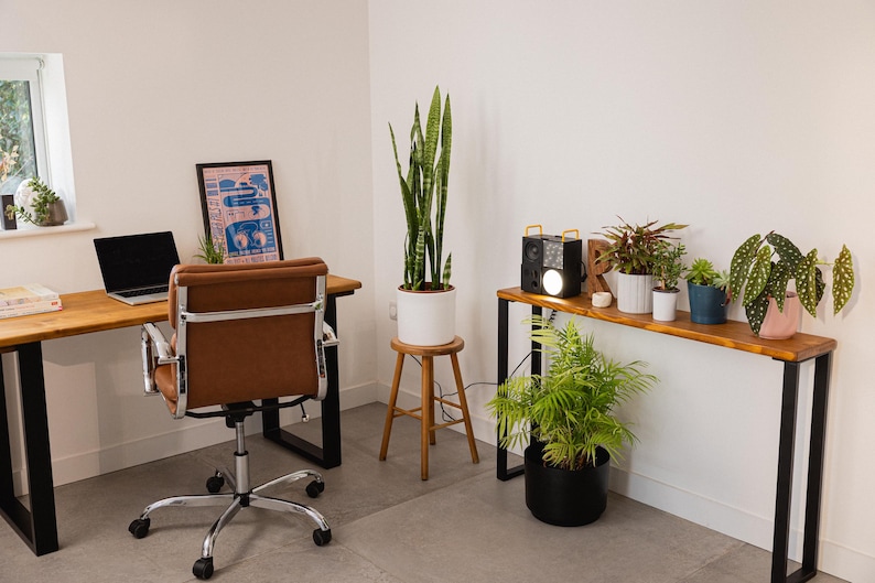 Slim wooden console table with black square metal legs, positioned over a radiator and decorated with potted houseplants and a speaker, styled in a modern workspace as a side desk with matching furniture.