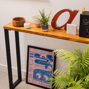 Close up shot of a wooden console table with black square metal legs, styled as a radiator shelf in a modern hallway with plants, books, and decorative objects on top.