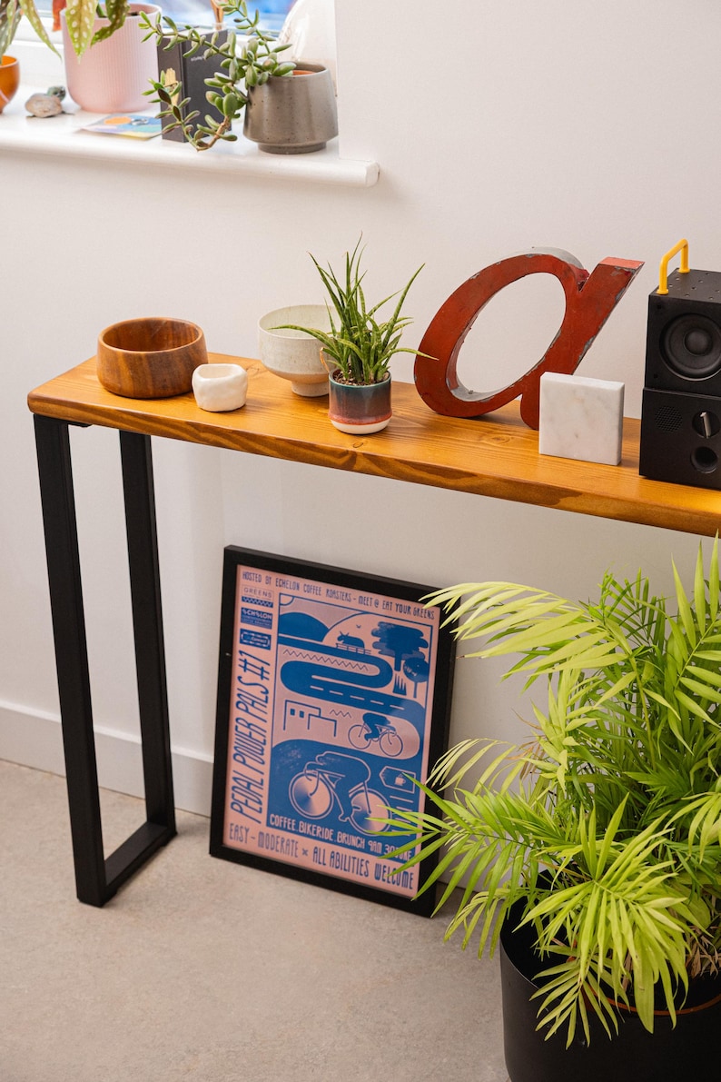 Close up shot of a wooden console table with black square metal legs, styled as a radiator shelf in a modern hallway with plants, books, and decorative objects on top.