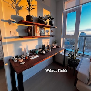 Simple solid wood desk with a dark wood finish and black steel legs. 2 wooden shelves above it, on the wall. Placed near a large window with views of the city.