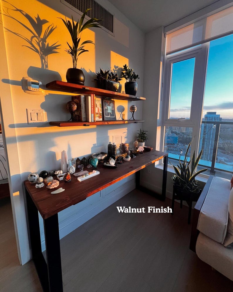 Simple solid wood desk with a dark wood finish and black steel legs. 2 wooden shelves above it, on the wall. Placed near a large window with views of the city.