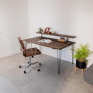 Wide shot of a minimalist workspace featuring a dark wood desk with black hairpin legs and a raised shelf. Styled with books, ceramics, and greenery, the setup is paired with a brown leather office chair on castors. Set in a bright room with exposed wooden beams, concrete-style flooring, and plenty of natural light.