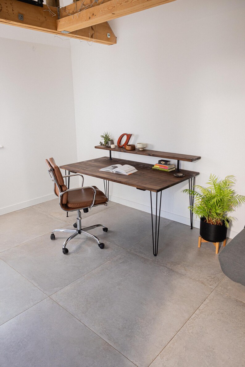 Wide shot of a minimalist workspace featuring a dark wood desk with black hairpin legs and a raised shelf. Styled with books, ceramics, and greenery, the setup is paired with a brown leather office chair on castors. Set in a bright room with exposed wooden beams, concrete-style flooring, and plenty of natural light.