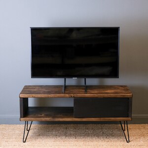 May include: A dark brown wooden TV stand with a black drawer and a shelf. The stand has hairpin legs and is supporting a black TV on a stand.